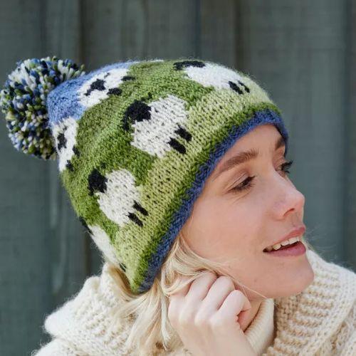 Woman wearing a green knitted hat with black and white patterns and a blue pom-pom against a gray background.