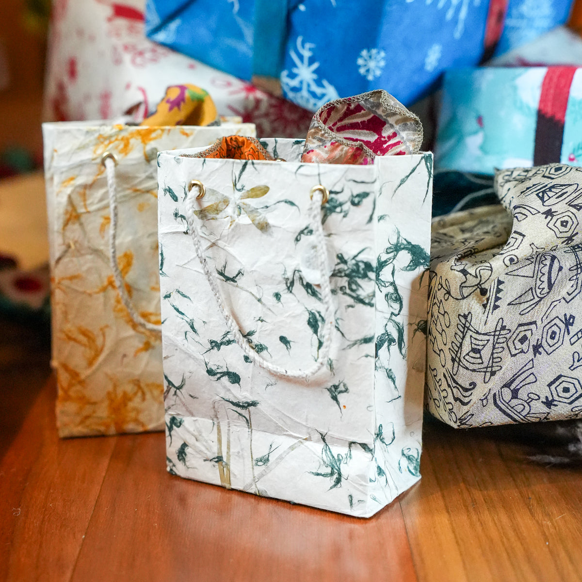 Three decorative paper gift bags with patterned wrapping paper and rope handles, arranged on a wooden surface with more wrapped packages blurred in the background.