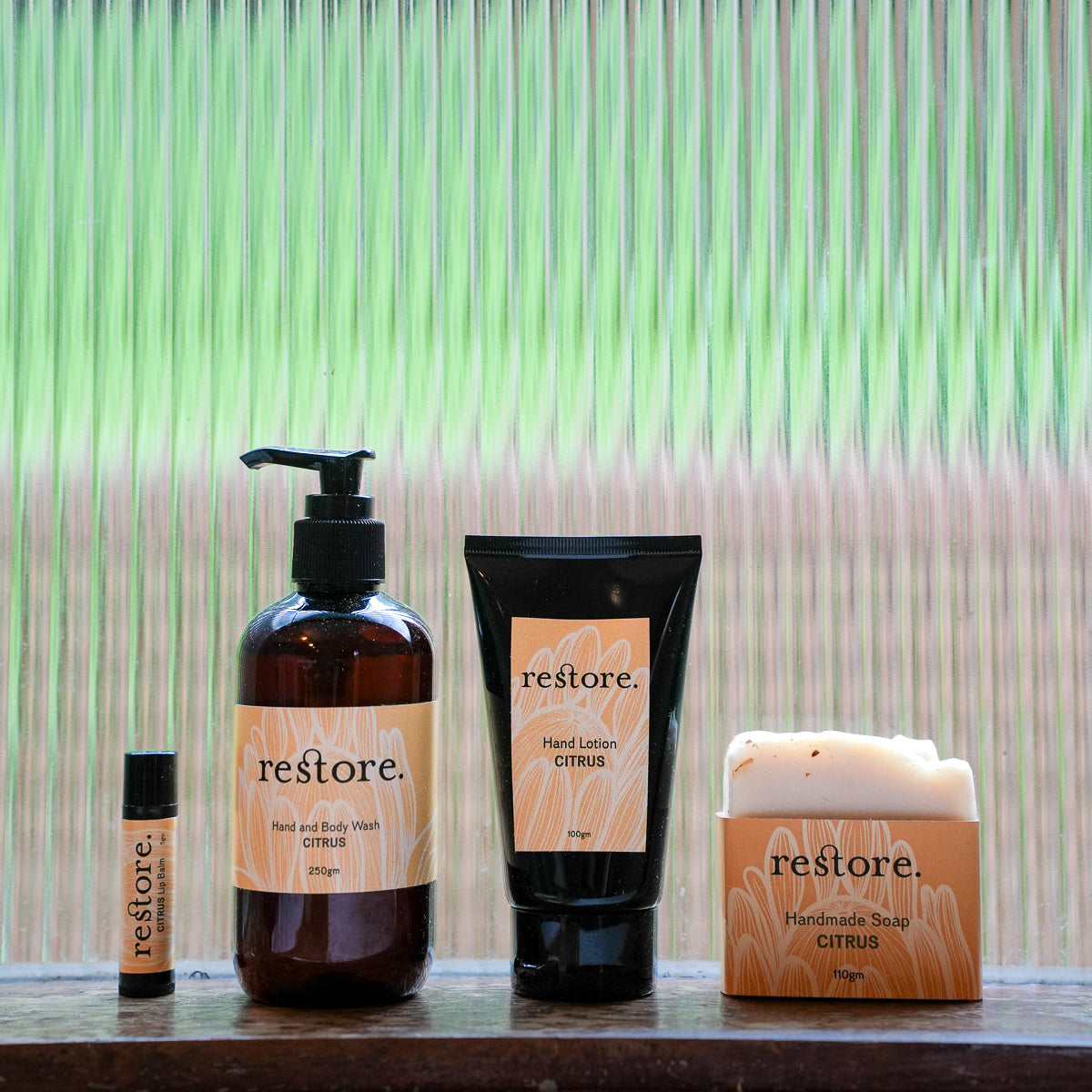 Four Restore brand bath and body products arranged on a wooden ledge in front of a corrugated glass window with green-tinted light: a small amber bottle, a large amber pump bottle, a black hand lemon citrus tube, and a white handmade soap bar in a wooden box.