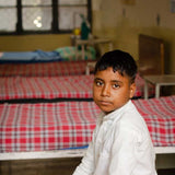 Child sitting on a bed with red and white checkered sheets in a room with other beds and furniture.