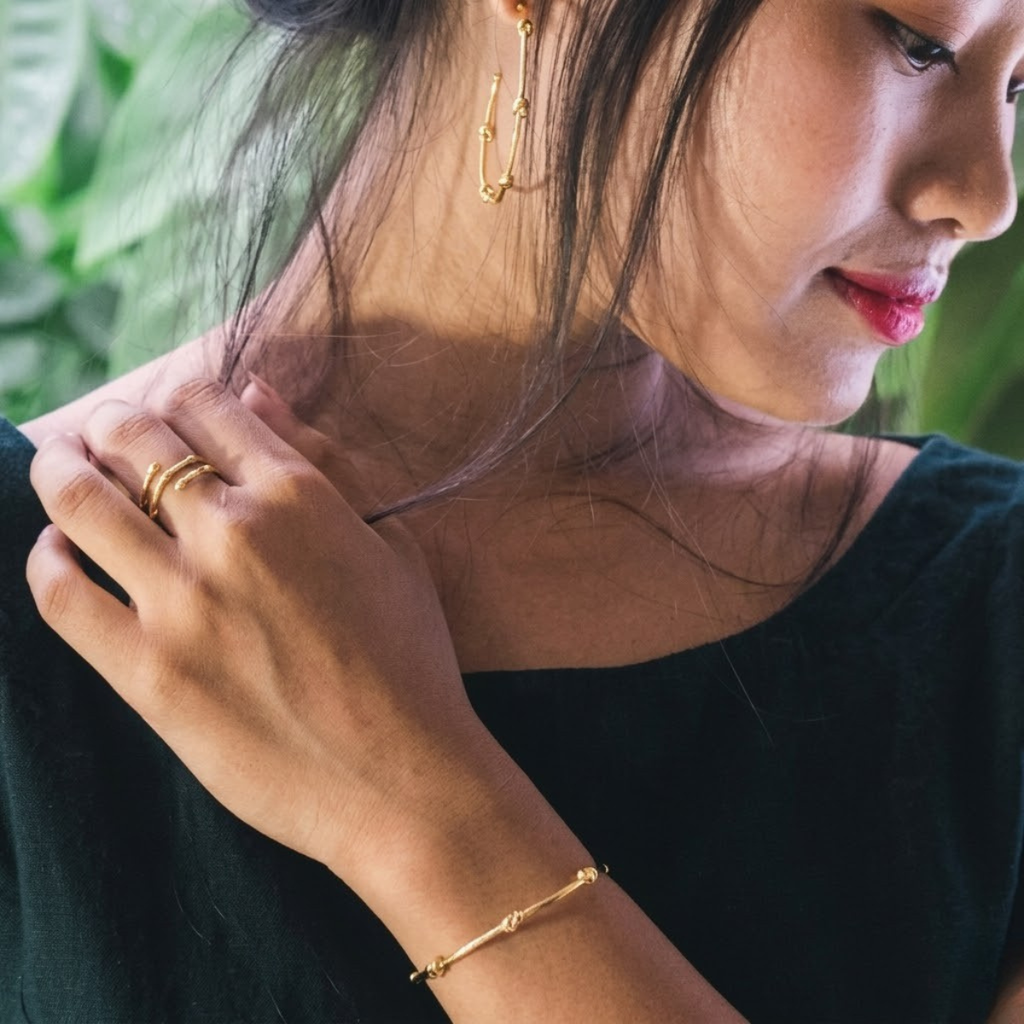Woman wearing gold jewelry including earrings, ring, and bracelet with a blurred green background
