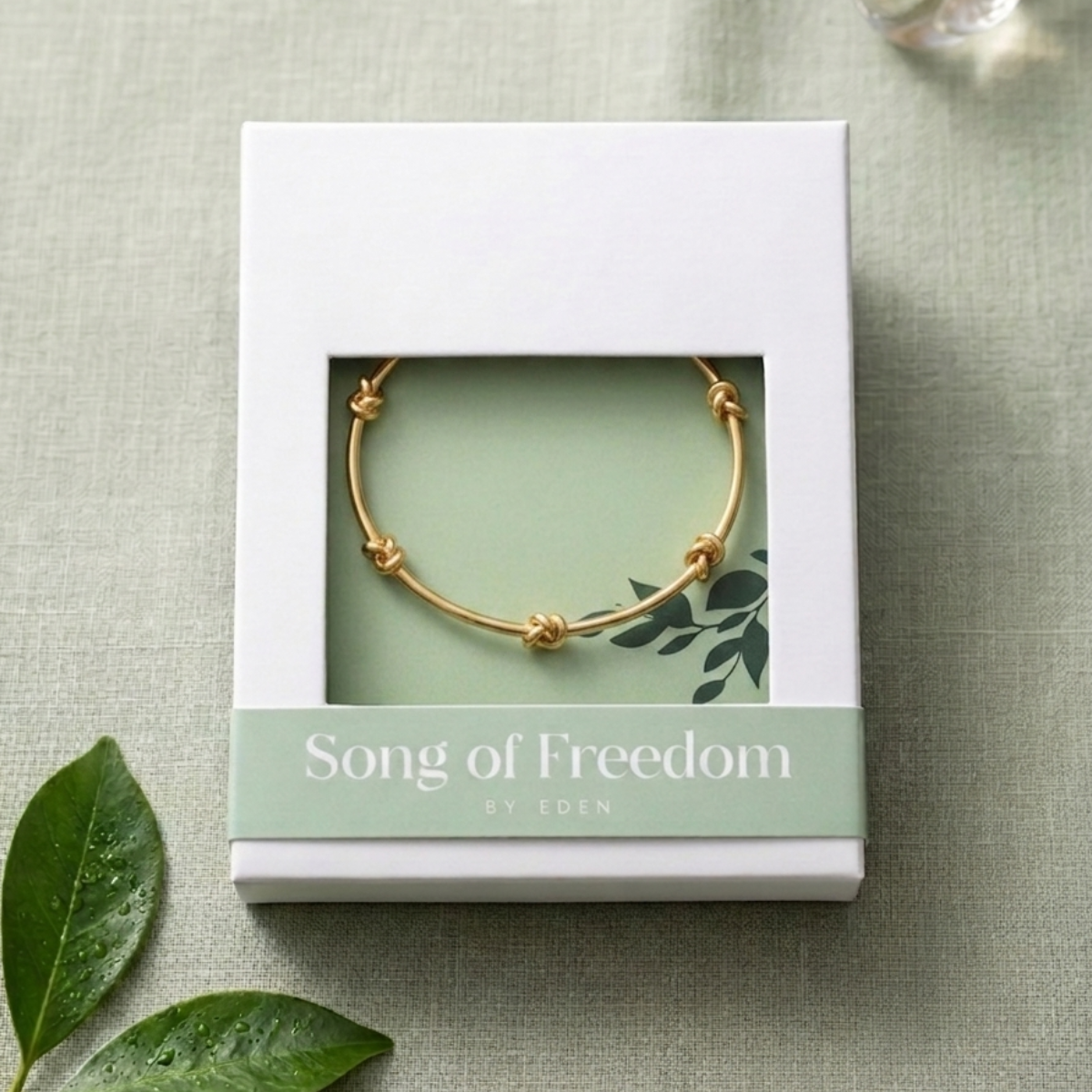 Gold bracelet in a 'Song of Freedom by Eden' box on a textured green surface with leaves.