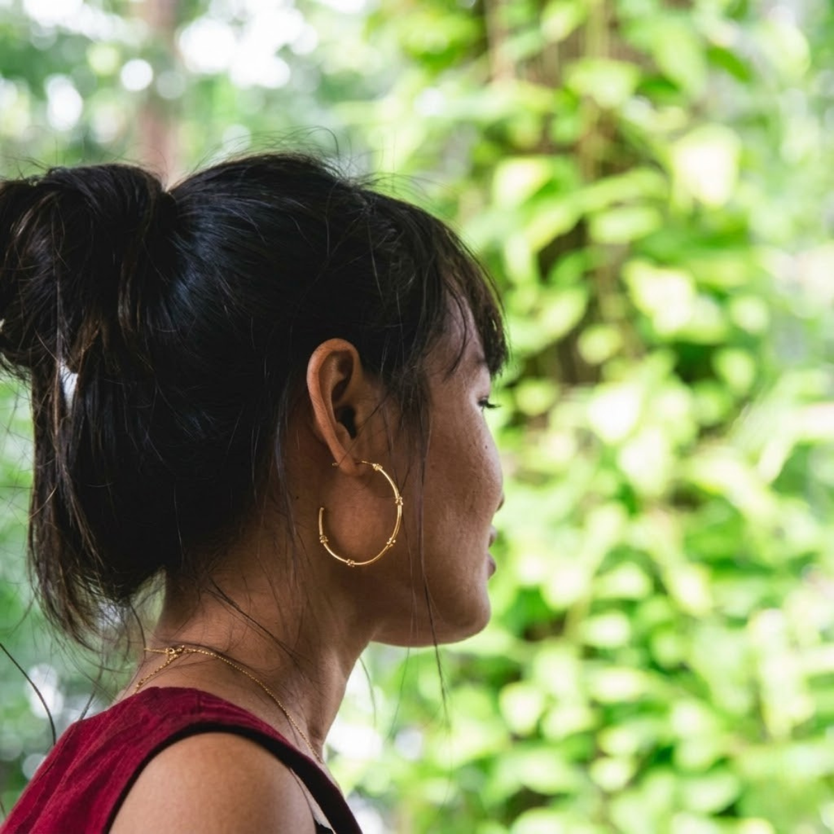 Woman with a ponytail and gold hoop earrings against a green leafy background